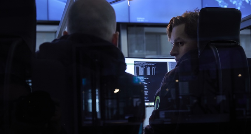 Zwei Personen sitzen in einem dunklen Raum vor Computermonitoren, die Codes anzeigen. Eine Person schaut die andere an, während das Licht des Bildschirms sie anstrahlt, was auf eine konzentrierte Arbeits- oder Cybersicherheitsumgebung schließen lässt.