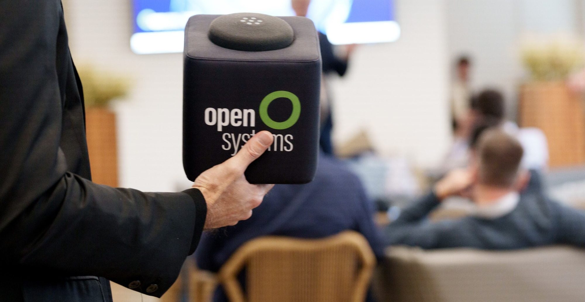 A person holds a black cube-shaped microphone with the “open systems” logo, while people sit and listen in a blurred conference or meeting setting in the background.