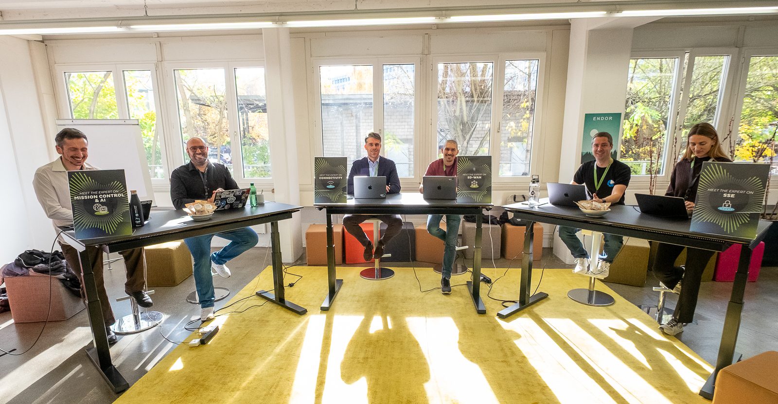 Sechs Personen sitzen an Stehpulten mit Laptops in einem hellen Raum mit großen Fenstern. Das Sonnenlicht strömt herein, und auf den Tischen stehen Snacks. Alle wirken engagiert und lächeln, was auf ein kollaboratives Arbeitsumfeld schließen lässt.