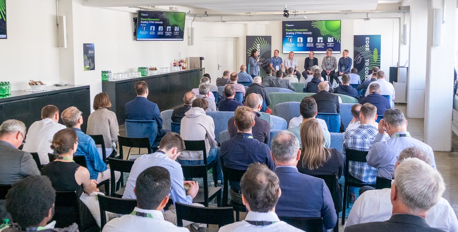 Eine Gruppe von Personen sitzt vor einem Podium mit fünf Rednern im vorderen Teil eines Konferenzraums, hinter ihnen befinden sich Bildschirme mit Informationen zum Podium und zur Veranstaltung. Auf den Seitentischen stehen Wasserflaschen bereit.