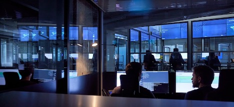 Menschen arbeiten an Computerarbeitsplätzen in einem dunklen Raum voller großer Monitore mit Daten und Karten - ein Command Center bzw. Security-Operations-Umfeld, betrieben mit Managed-Connectivity-Services.