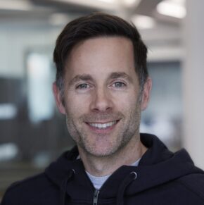 A man with short brown hair and light stubble smiles at the camera. He is wearing a dark zip-up hoodie and is standing indoors, with a blurred office background at the NIS-2 Congress.