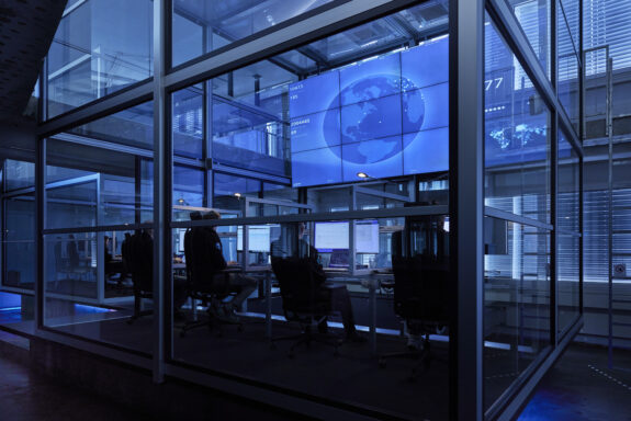 A modern control room with glass walls, several Mikron SASE customer teams working at computer desks, and large digital screens displaying a world map and data in blue tones.