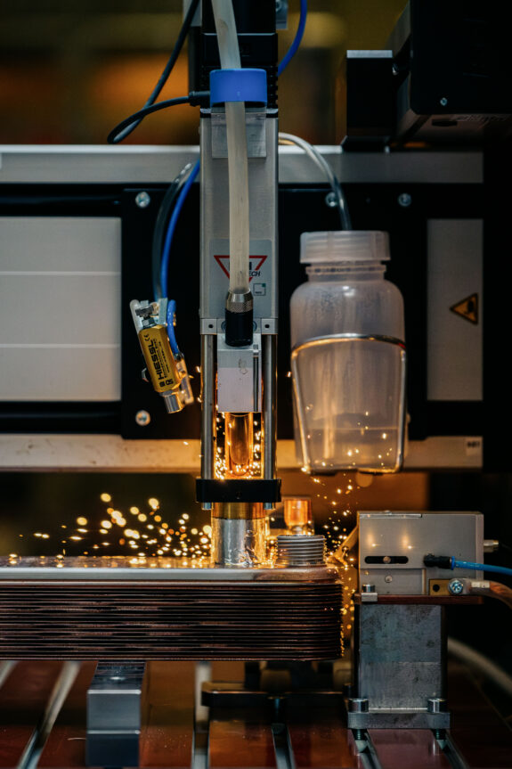 A close-up of a Kelvion robotic machine welding metal parts, with bright sparks flying around, surrounded by industrial components and a clear plastic container attached to the equipment.