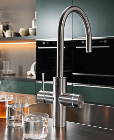 A modern stainless steel kitchen faucet with a tall, curved spout stands on a countertop, next to three glasses filled with water in a stylish kitchen with dark cabinets and built-in ovens in the background.