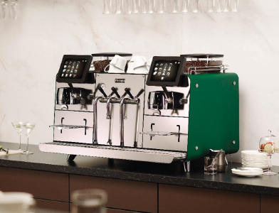 A modern commercial Kelvion espresso machine with dual touch screens and green side panels sits on a countertop, surrounded by cups, glasses, and coffee accessories in a stylish kitchen or café setting.