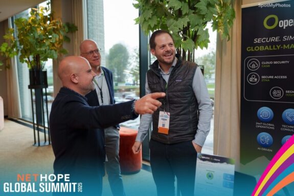 Three men in business attire stand talking and smiling near a conference booth at the NET HOPE Global Summit 2023, discussing NGOs and security. One man points forward as the others listen and laugh, with lush green plants and large windows behind them.