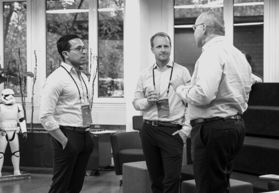 Three men in business attire stand indoors, engaged in conversation. Two of them hold drinks. They wear conference badges. In the background, there is a life-size Stormtrooper figure and large windows letting in daylight.