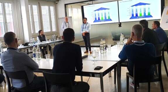 A group of people sit around tables in a bright conference room, listening to two men presenting graphs displayed on large screens at the front of the room. Bottles of water and glasses are on the tables.