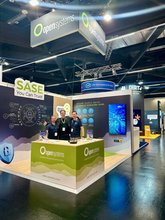Three people stand behind a booth at a tech conference, with “Open Systems” branding and “SASE You Can Trust” displayed above. The booth features monitors, brochures, and a large circular hanging sign overhead.