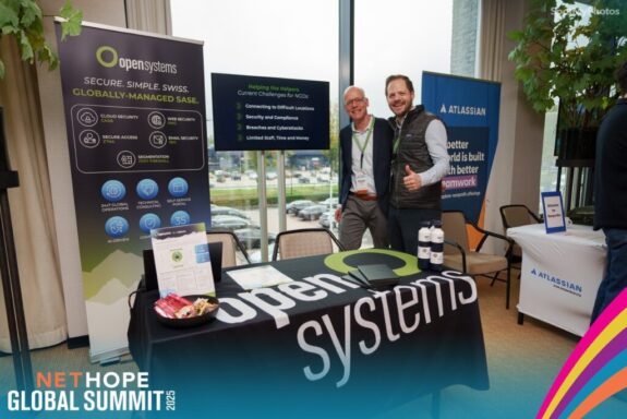 Two smiling people stand behind an Open Systems booth at a conference, highlighting NGOs and security. Banners, a branded tablecloth, water bottles, and promo items are visible, with an Atlassian banner nearby. "NetHope Global Summit 2023" is shown below.