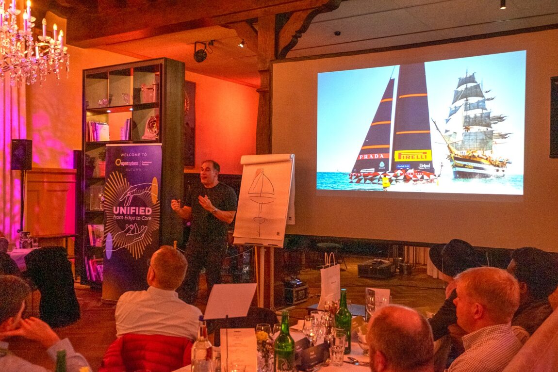A man gives a presentation to an audience in a warmly-lit room, with a screen showing images of a racing sailboat and a tall ship. There are drinks on tables and a flip chart with a boat sketch.