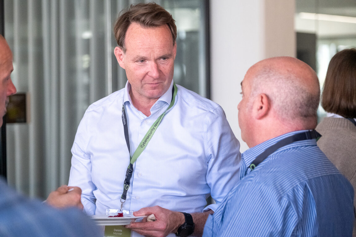 A man in a light blue shirt wearing a lanyard listens attentively to another man in a striped shirt at an indoor event. There are other people and glass walls in the background.