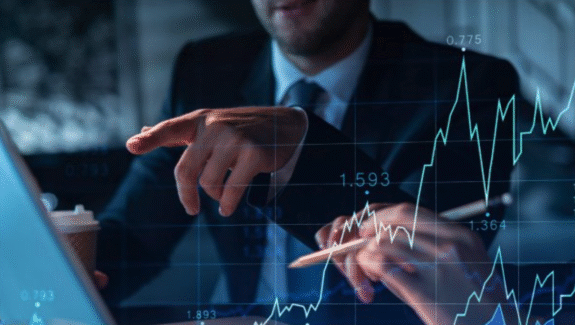 Two people in business attire discuss financial data, pointing at a laptop screen. Transparent graphs and stock market numbers are overlaid on the image, indicating financial analysis or trading.